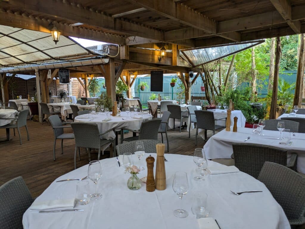 Terrasse de restaurant couverte en bois, tables dressées en blanc, chaises en osier. Verdure luxuriante, lanternes et menu COTE 29.5€.