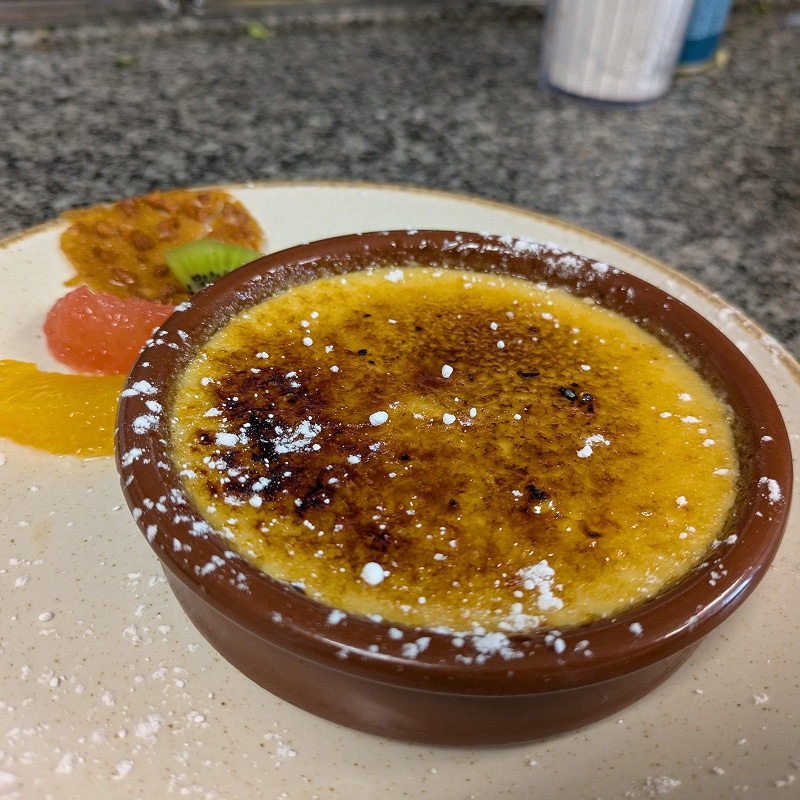 Crème brûlée dorée avec sucre glace dans ramequin brun, garnie de fruits (orange, pamplemousse, kiwi) sur assiette claire.