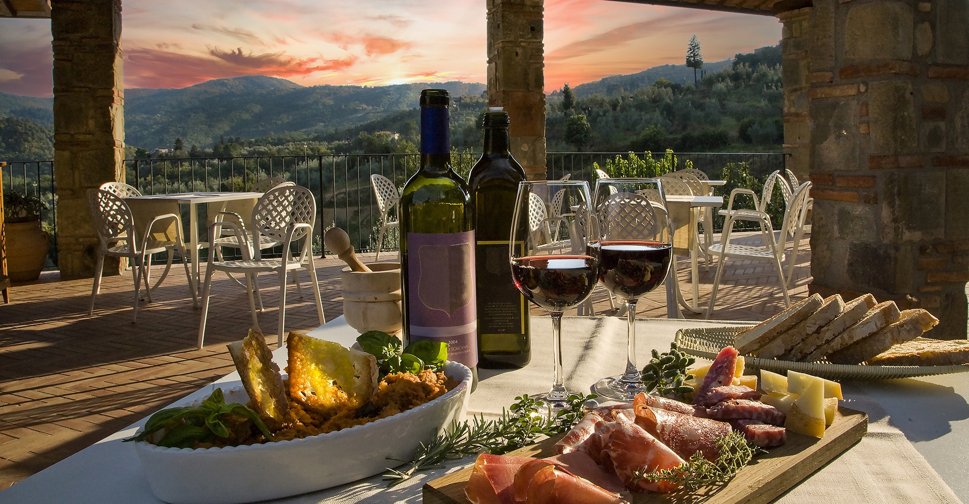 Terrasse toscane au coucher du soleil avec table dressée: vin rouge (2004 Toscana), charcuterie, fromage, bruschetta. Vue sur collines.