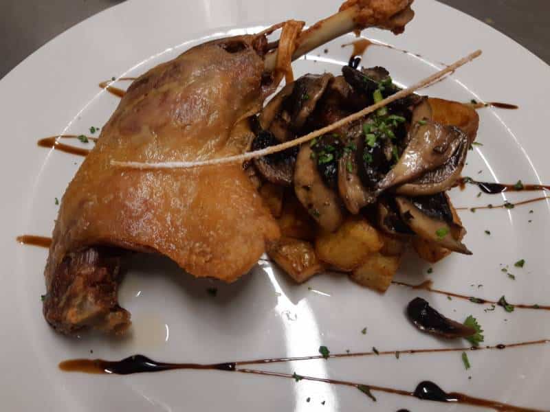Cuisse de canard confite dorée avec peau croustillante, accompagnée de pommes de terre rissolées et champignons de Paris poêlés, sauce au jus.