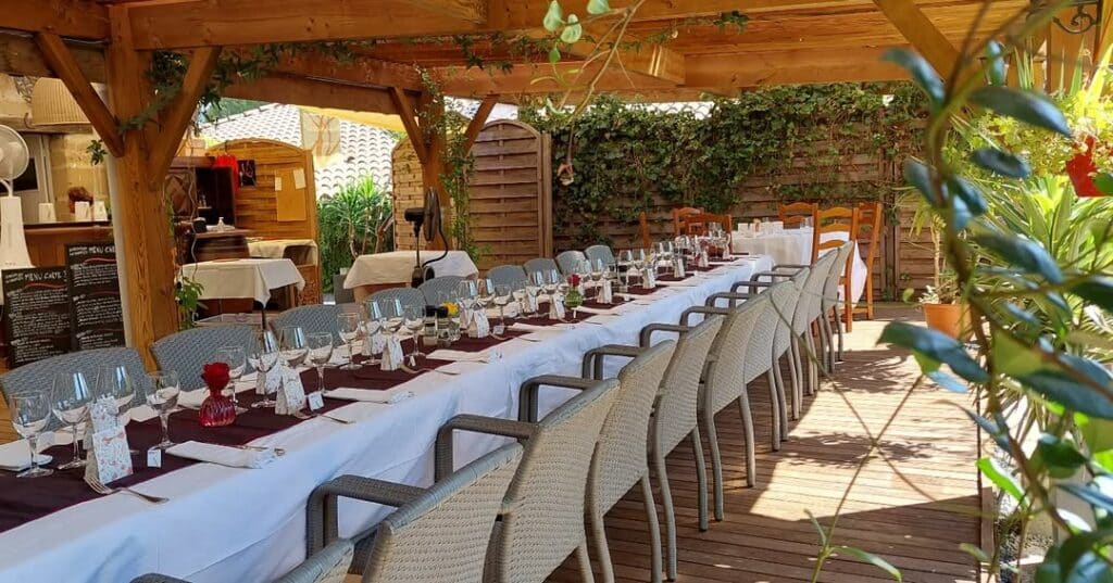 Longue table dressée sous une pergola en bois pour un repas extérieur. Nappe blanche, chemin bordeaux, verres. Panneaux 'MENU CARTE'.